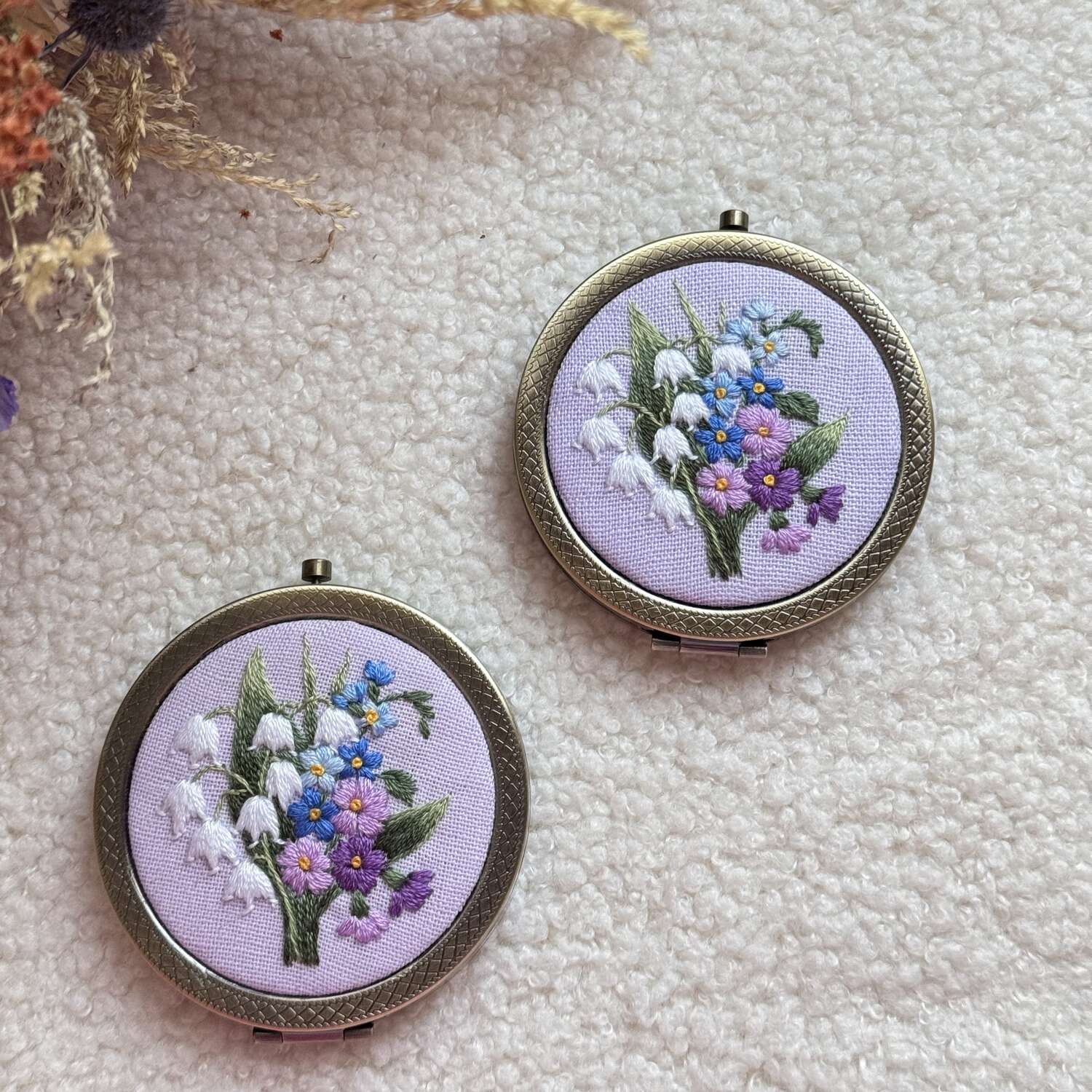 Käsin kirjottu taskupeili, kielo ja lemmikki -aiheinen kuvio kannessa