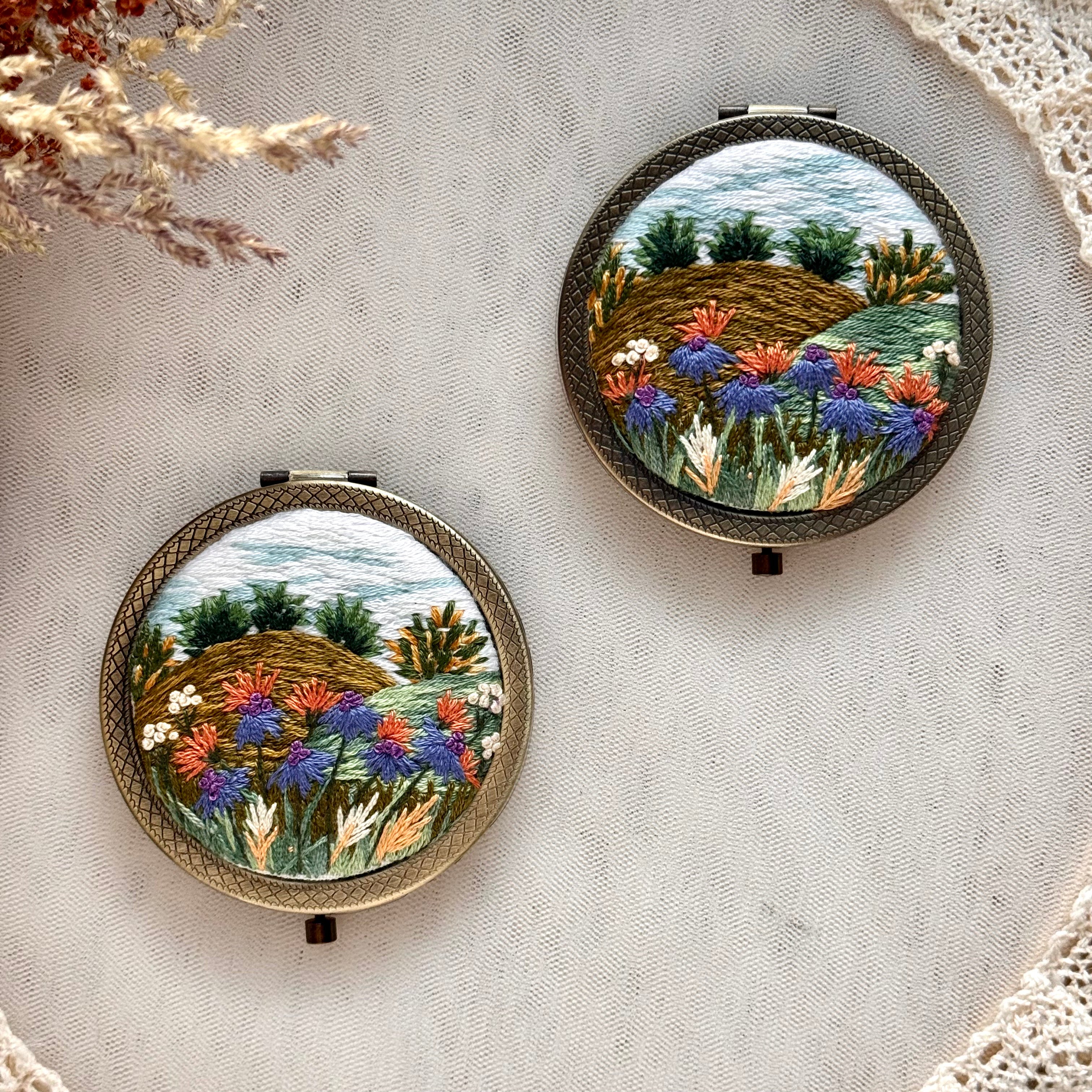 Käsin kirjottu taskupeili, kukkamäki-aiheinen kuvio kannessa