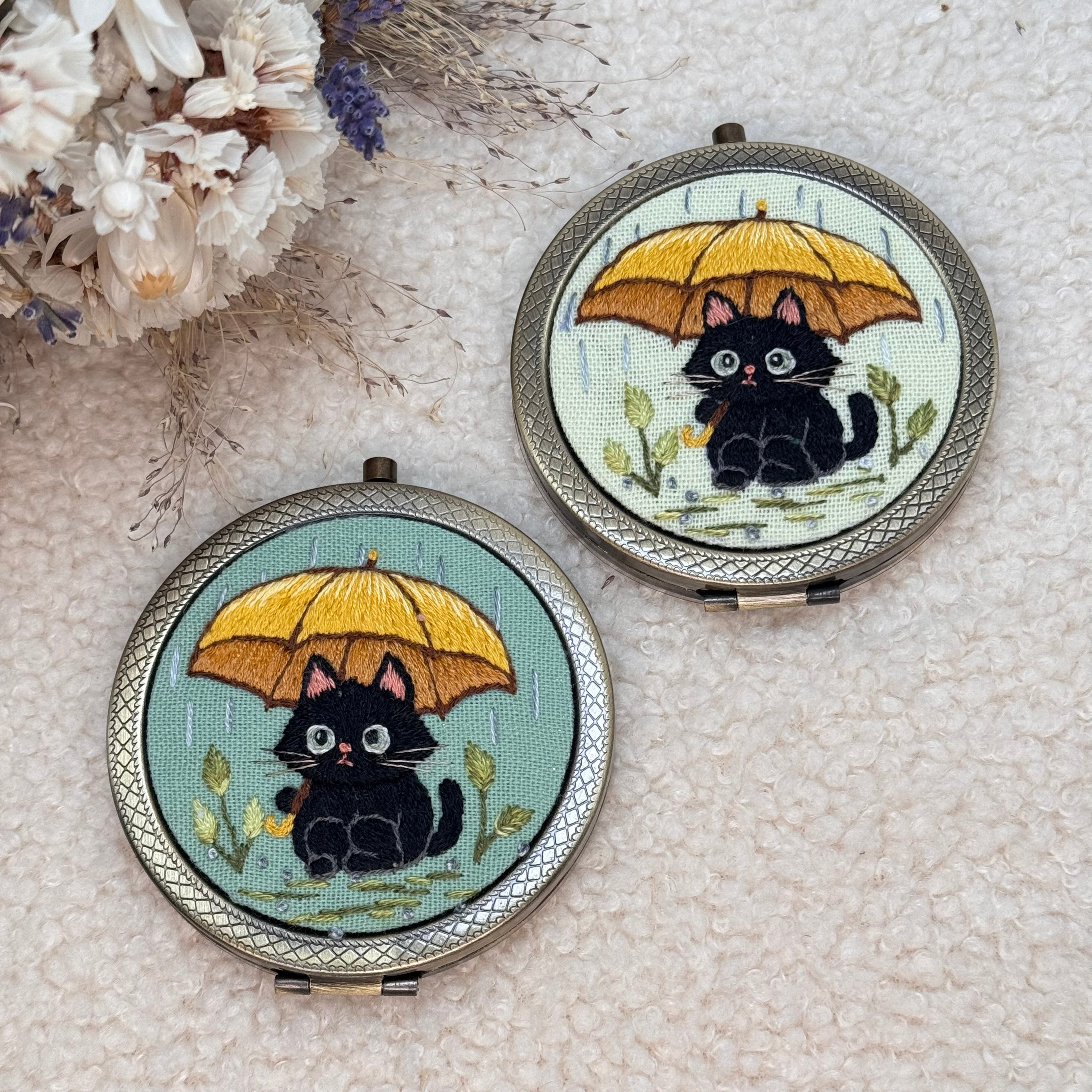 Käsin kirjottu taskupeili, musta kissa ja keltainen sateenvarjo -aiheinen kuvio kannessa