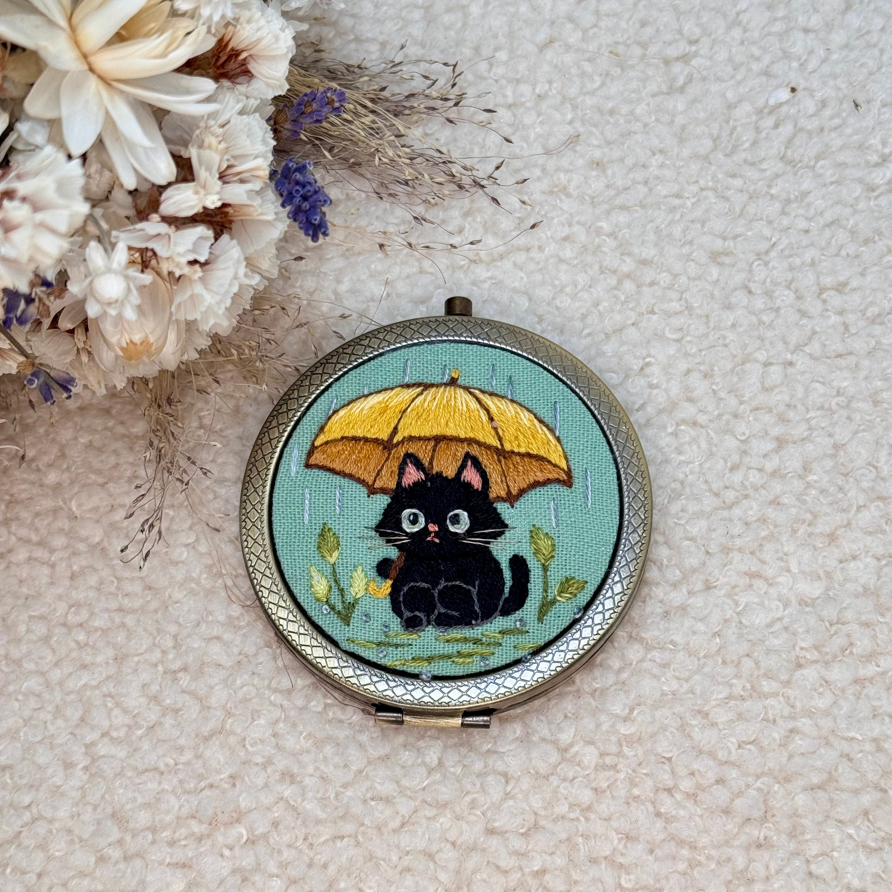 Käsin kirjottu taskupeili, musta kissa ja keltainen sateenvarjo -aiheinen kuvio kannessa