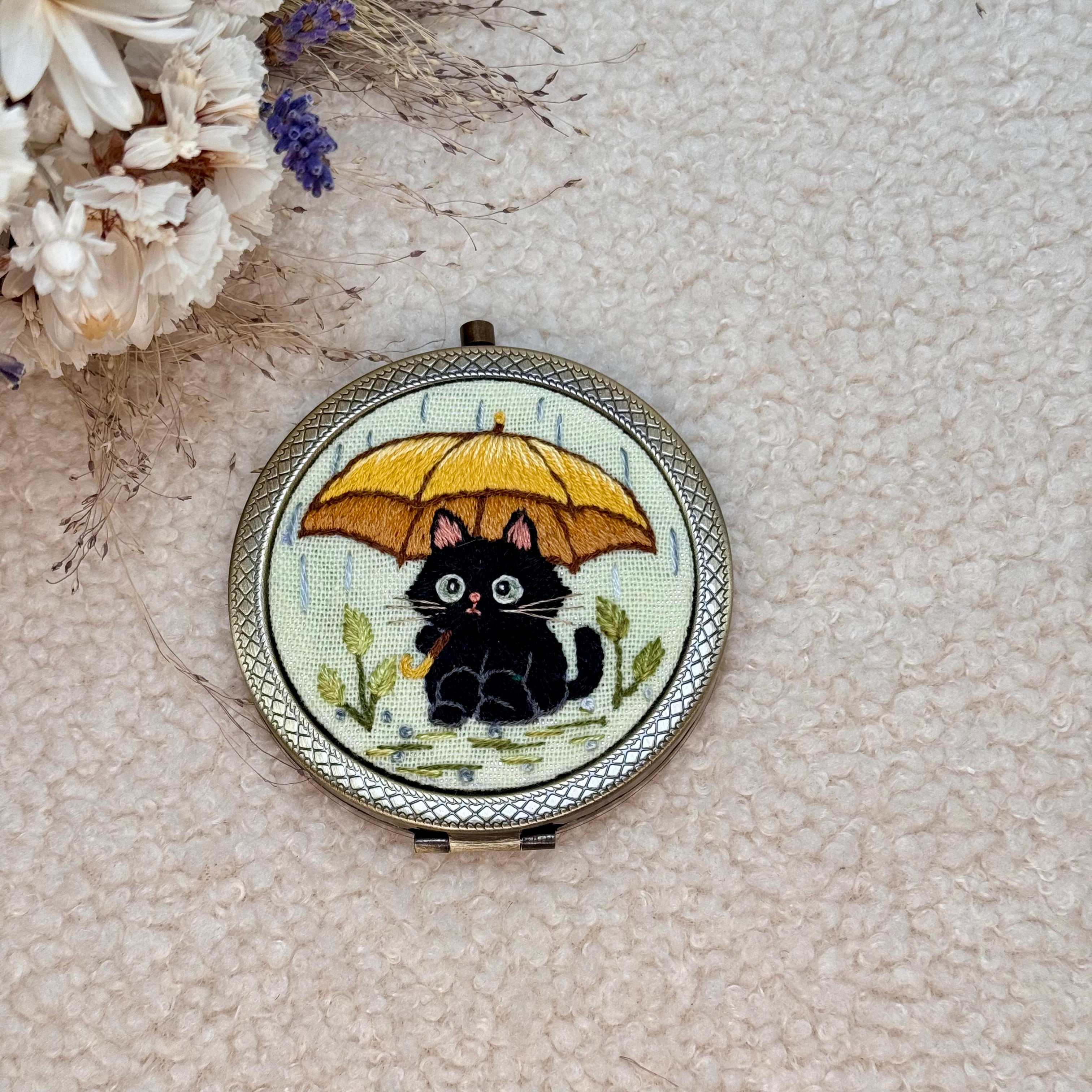 Käsin kirjottu taskupeili, musta kissa ja keltainen sateenvarjo -aiheinen kuvio kannessa