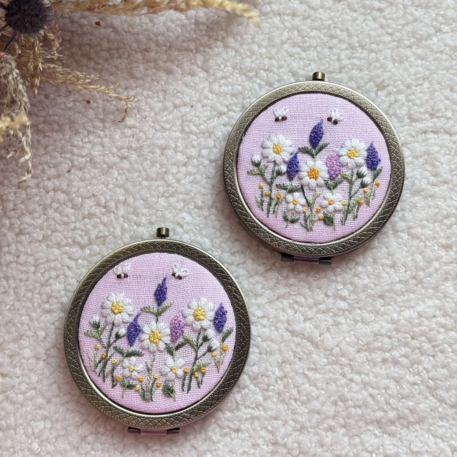 Käsin kirjottu taskupeili, syreeni ja päivänkakkara -aiheinen kuvio kannessa